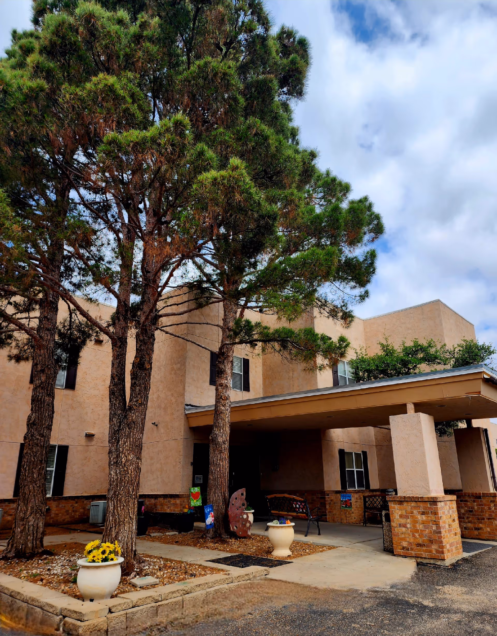 Exterior view of a beige stucco building with a covered entrance supported by brick pillars. There are tall pine trees and potted plants with flowers near the entrance. The sky is partly cloudy.