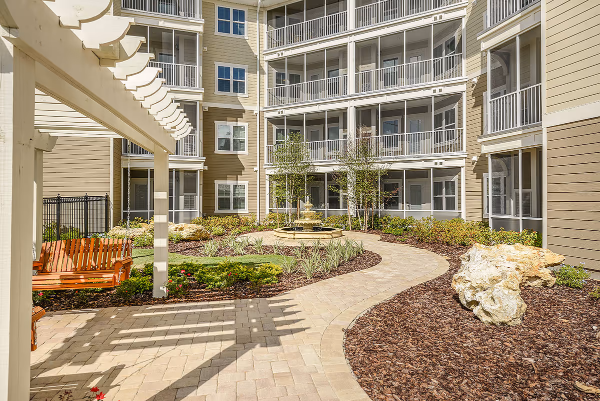Sunny landscaped courtyard of a multi-story apartment building with a paved walkway, fountain, pergola and a wooden swing.
