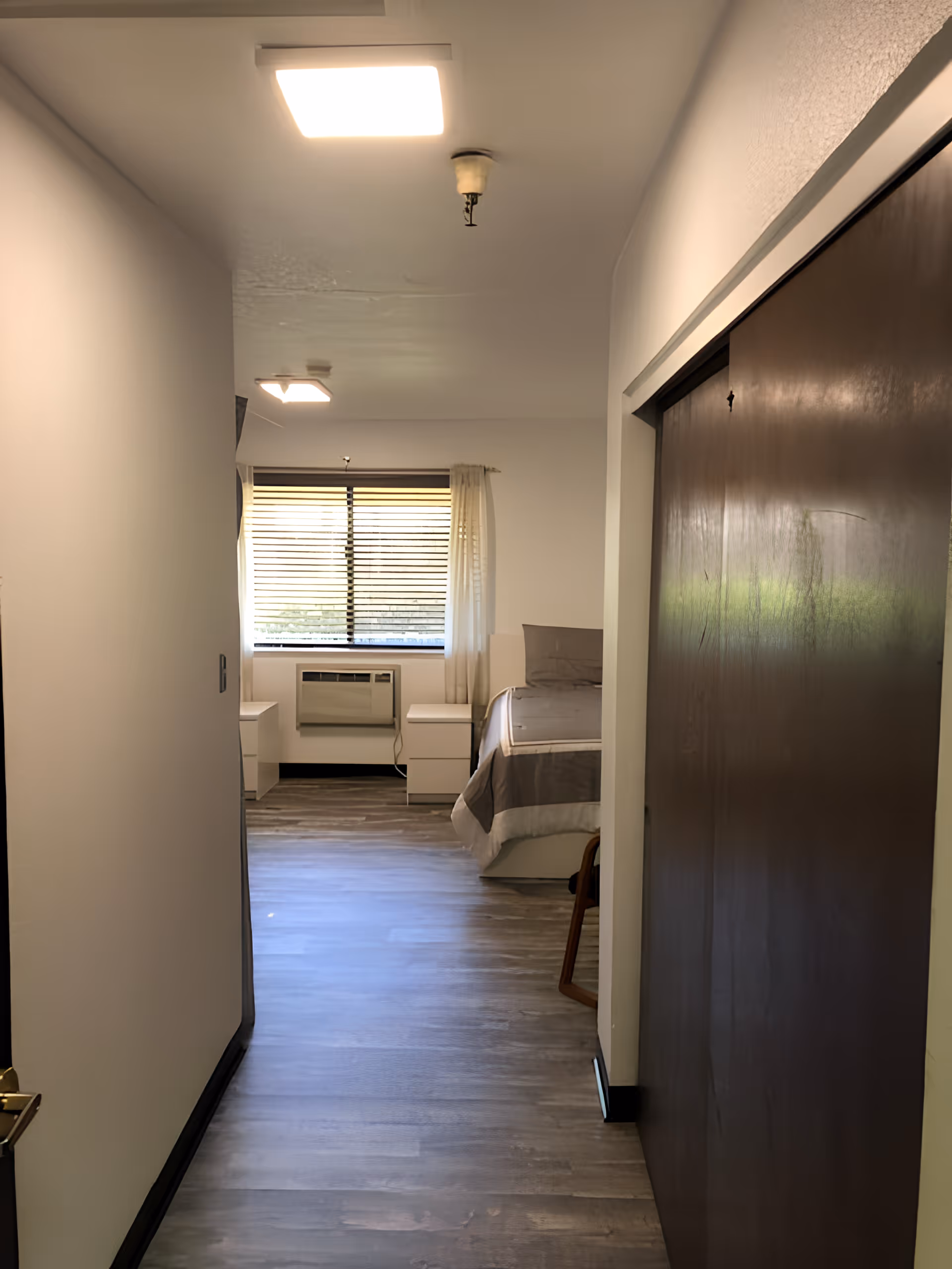 View down a hallway leading to a bedroom with a bed, two white nightstands, a window with blinds and sheer curtains, and an air conditioning unit below the window. The floor is wood-style laminate, and there is a large dark wooden sliding door on the right side of the hallway.