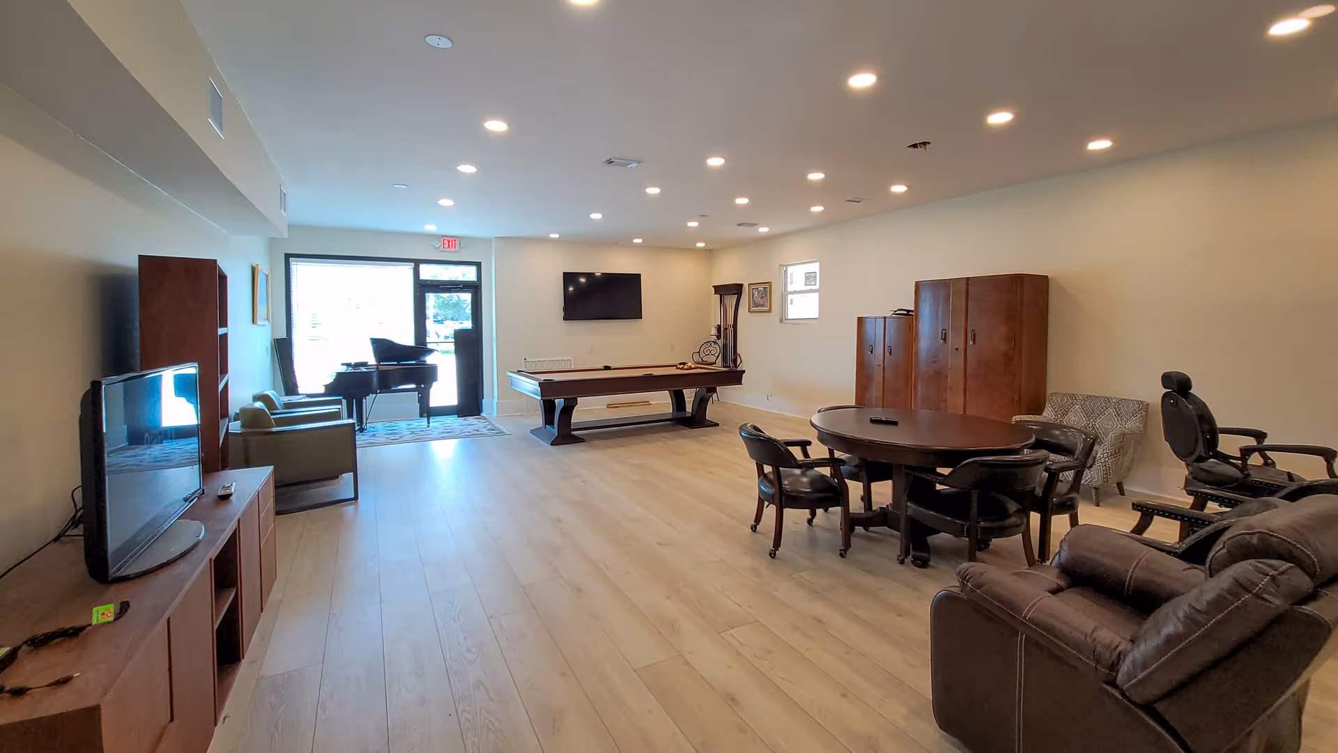 A spacious common area with light wood flooring and recessed ceiling lights. The room features a pool table, a round table with chairs, a flat-screen TV mounted on the wall, a piano near the glass door entrance, and various seating options including armchairs and recliners. There are wooden cabinets and a small window on the right wall.