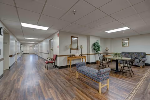 A long hallway in a senior living facility with wood-look flooring and white walls. On the right side, there is a seating area with a floral-patterned bench, a small table with chairs, a couch, and decorative items including lamps, mirrors, and a potted plant. The ceiling has recessed lighting panels.