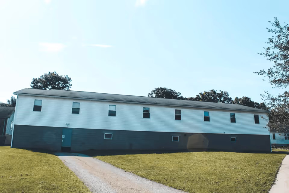 Exterior view of a long, single-story building with white siding on the upper half and gray concrete blocks on the lower half. The building has several windows and a green door at the front, with a driveway leading up to it. There is a grassy lawn surrounding the building and trees in the background under a clear blue sky.