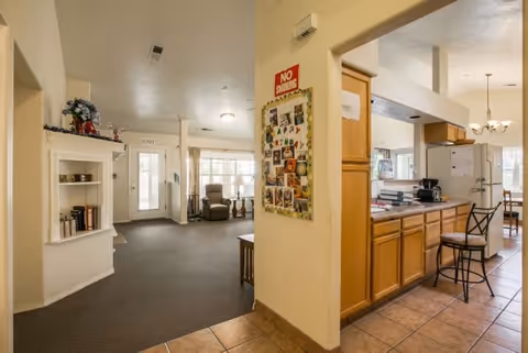 Interior view of a senior living facility showing a hallway leading to a sitting area with chairs near large windows. To the right, there is a kitchen area with wooden cabinets, a refrigerator, a coffee maker, and a bar stool. A bulletin board with photos and a 'No Smoking' sign is mounted on the wall between the hallway and kitchen.