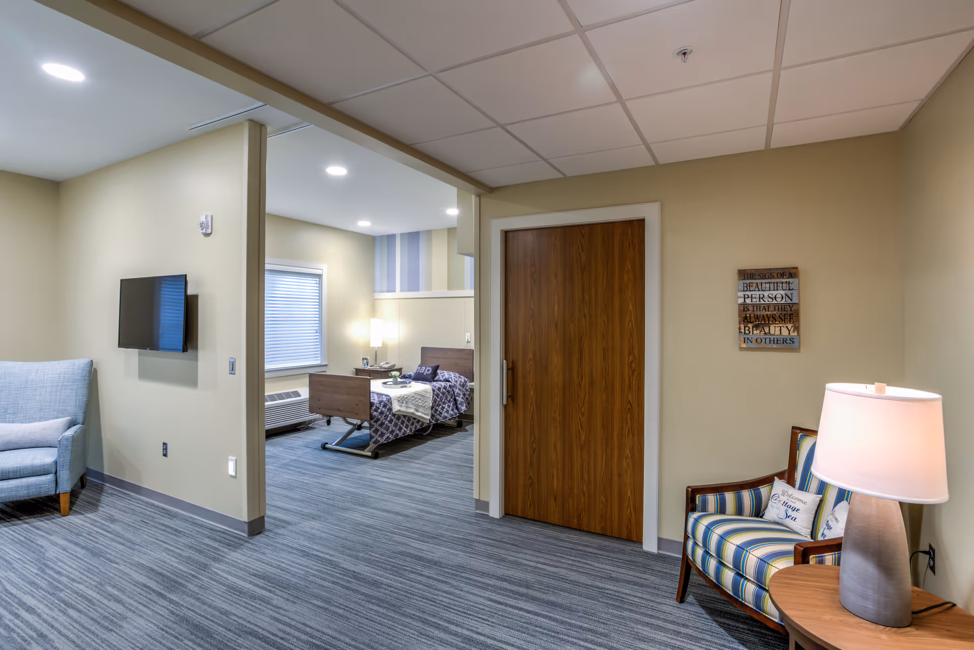 Interior view of a nursing facility room showing a sitting area with a striped armchair and lamp, a wall-mounted TV, and a hospital bed in an adjacent room with a window and bedside table.