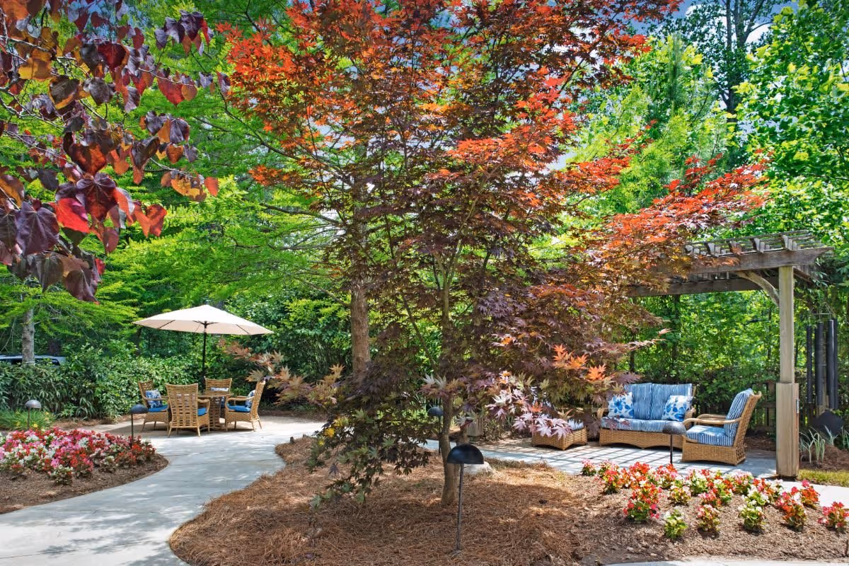 A peaceful outdoor garden area with a curved concrete pathway, colorful flower beds, and various trees with green and red leaves. There is a seating area with a table, chairs, and a large white umbrella on the left side, and a shaded wicker seating set with blue and white cushions under a wooden pergola on the right side.