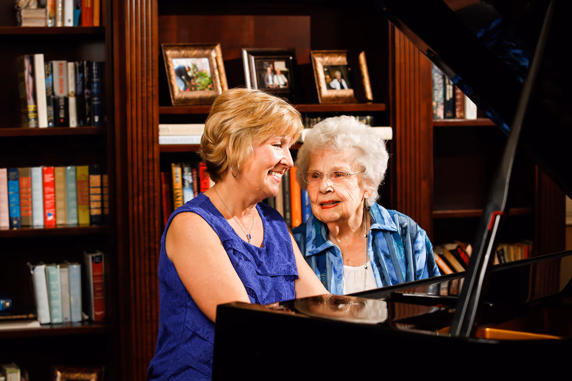 Two women, one elderly and one middle-aged, sitting together at a grand piano in a cozy room with wooden bookshelves filled with books and framed photos in the background. They appear to be enjoying a moment of music and companionship.