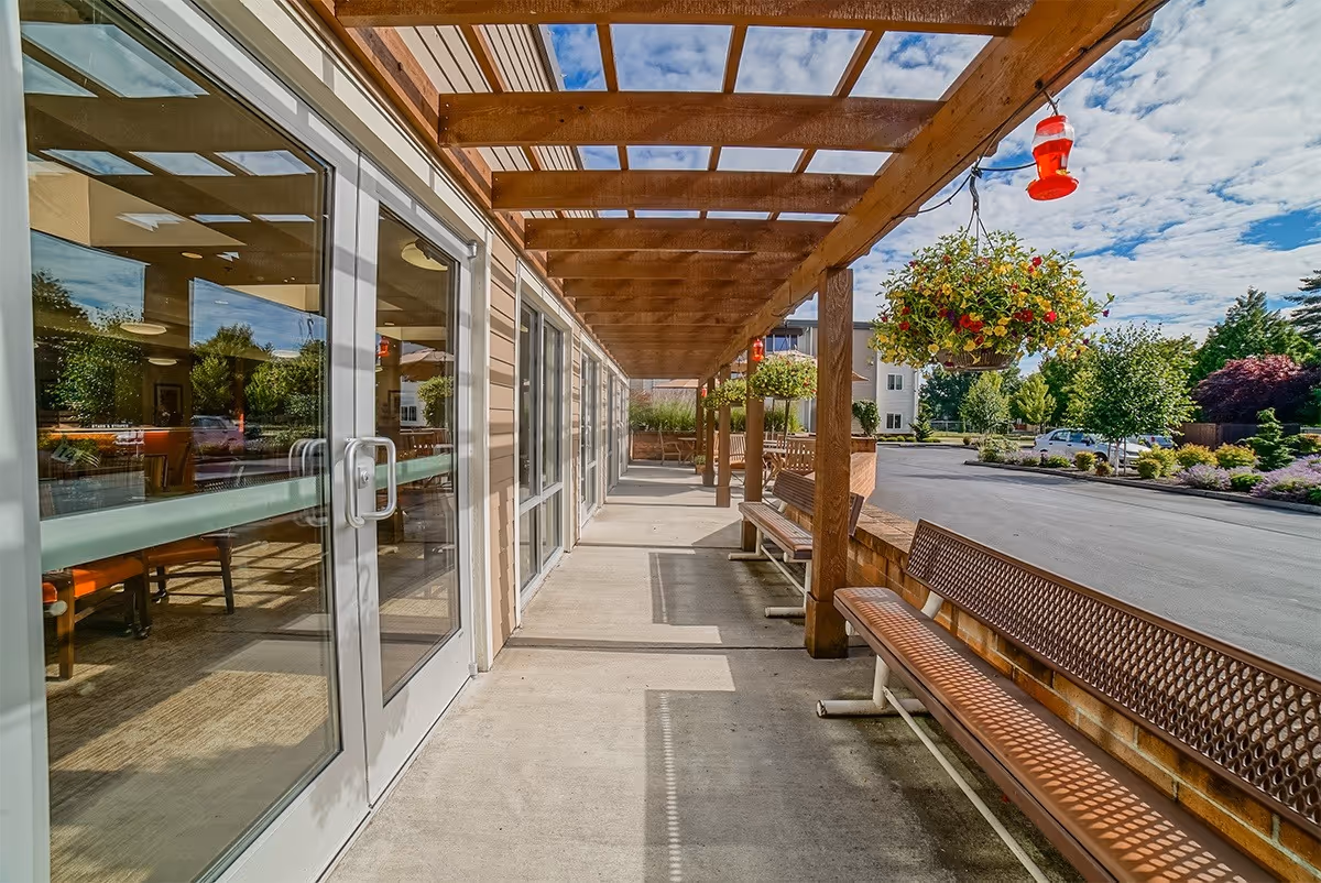 Outdoor covered walkway with wooden pergola overhead, hanging flower baskets, and metal benches along the side. Glass doors on the left lead inside a building, with a view of tables and chairs through the windows. The area is well-lit with natural sunlight and a partly cloudy sky is visible.