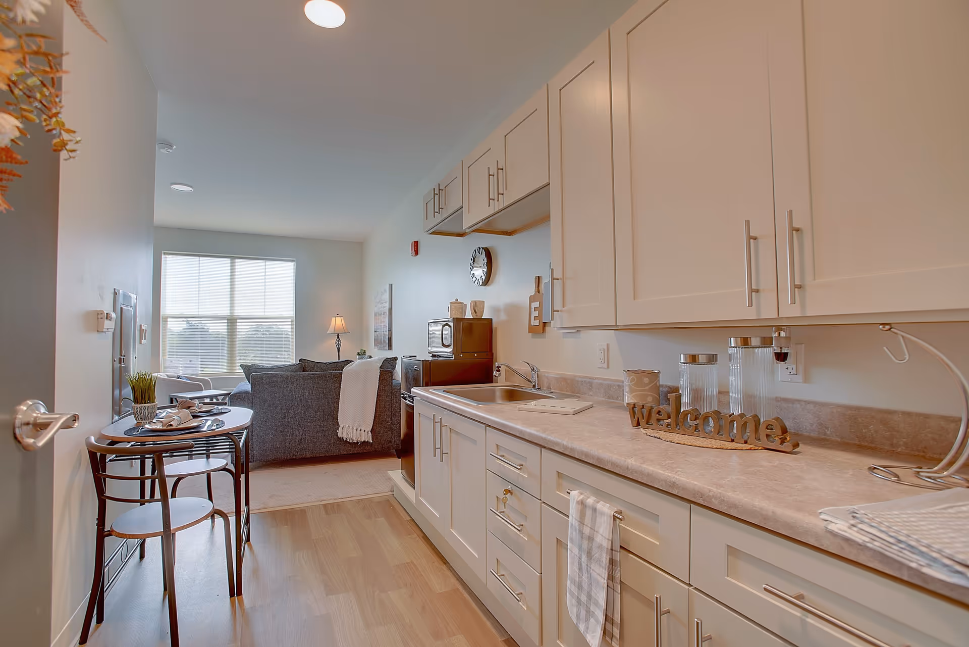 Open kitchenette with white cabinets and a long countertop overlooking a small dining table and a living room with a sofa and large window.