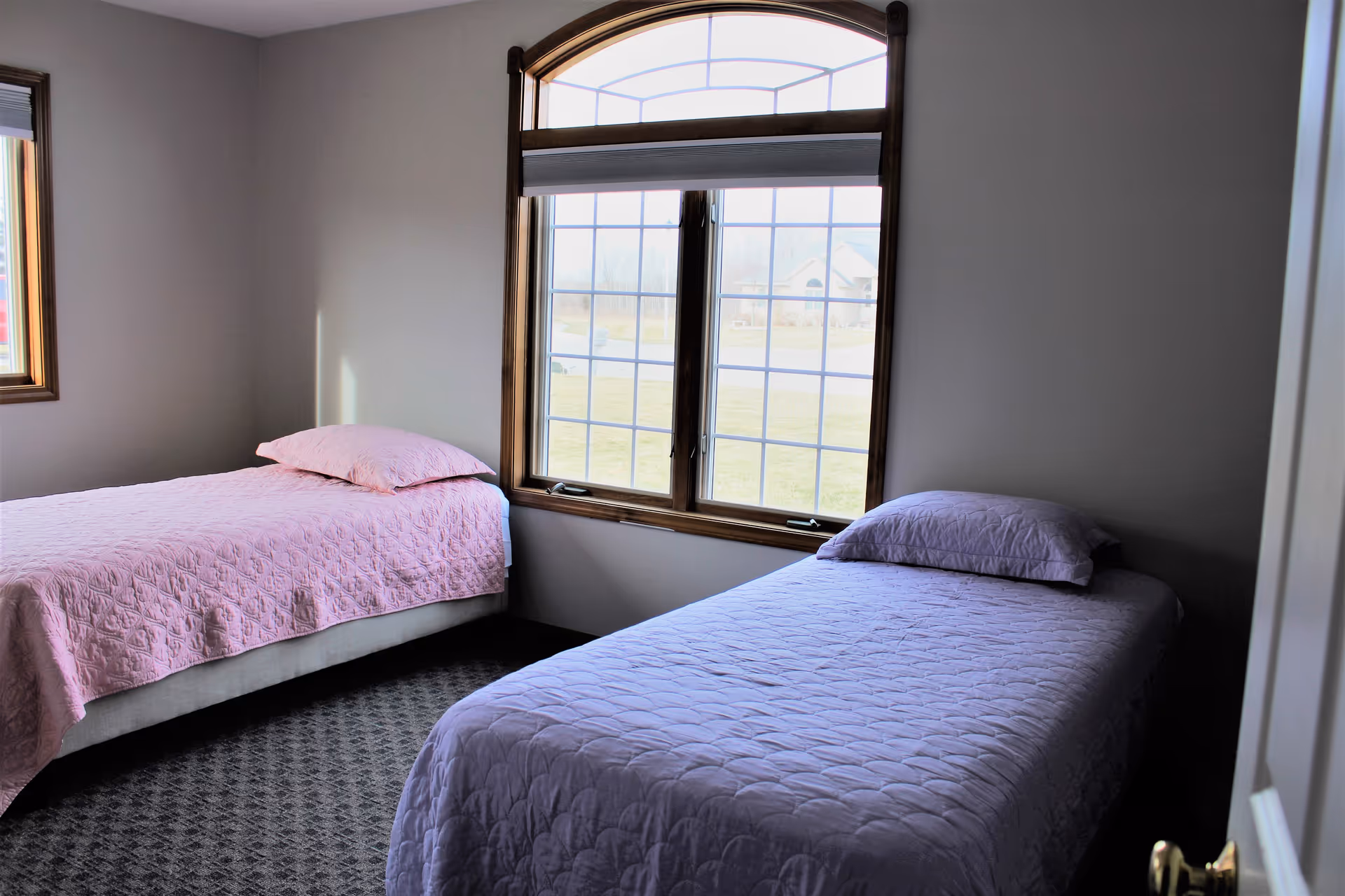 A bedroom with two single beds, one with a pink quilt and pillow and the other with a purple quilt and pillow. There is a large window with wooden trim between the beds, letting in natural light and showing a view of a grassy area and a house outside.
