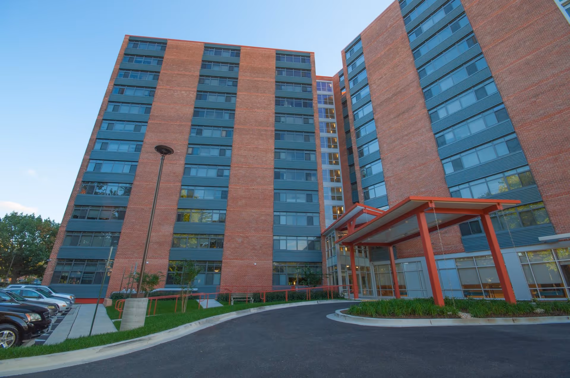 Exterior view of a multi-story brick apartment building with large windows and a covered entrance with red beams. There is a parking area with cars on the left side and some greenery around the entrance.