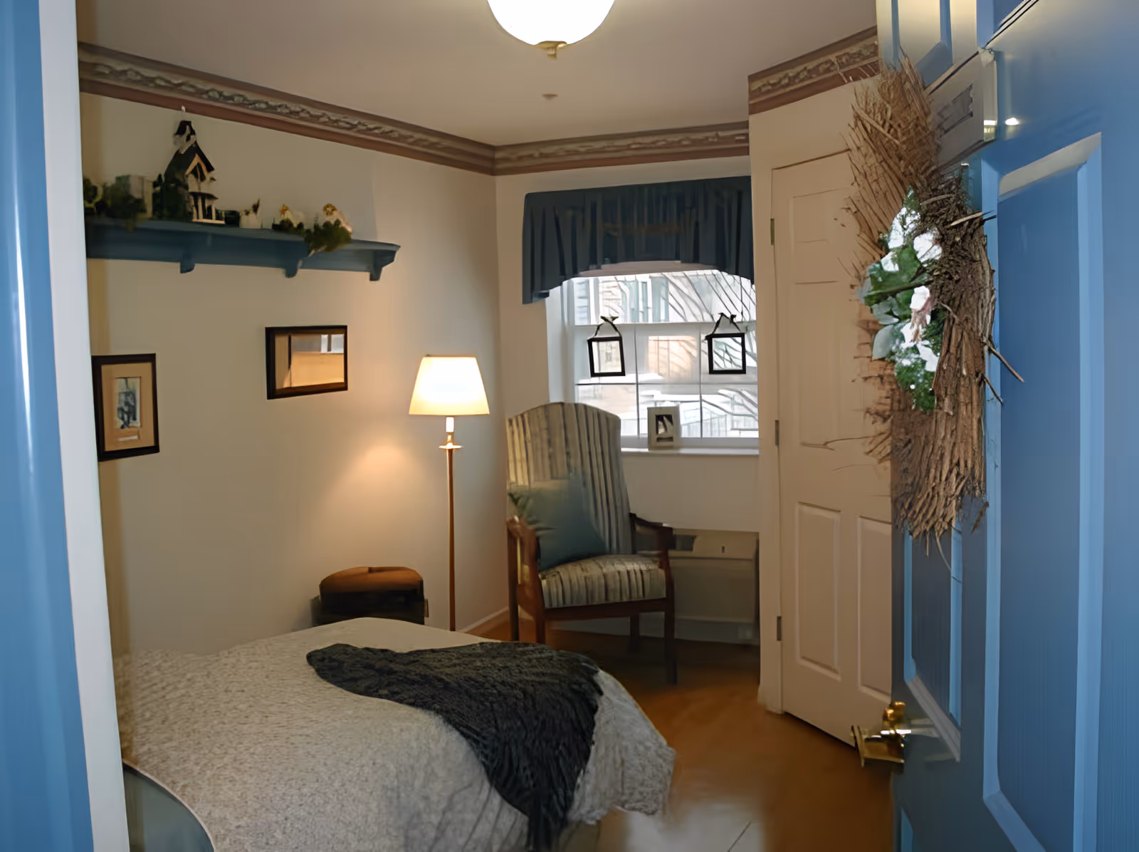 A cozy bedroom with a single bed covered in a light-colored quilt and a dark throw blanket. There is a wooden chair with a cushion near a window with blue curtains. A floor lamp is turned on next to the bed, and a shelf with decorative items is mounted on the wall above. The door is painted blue and has a wreath hanging on it.