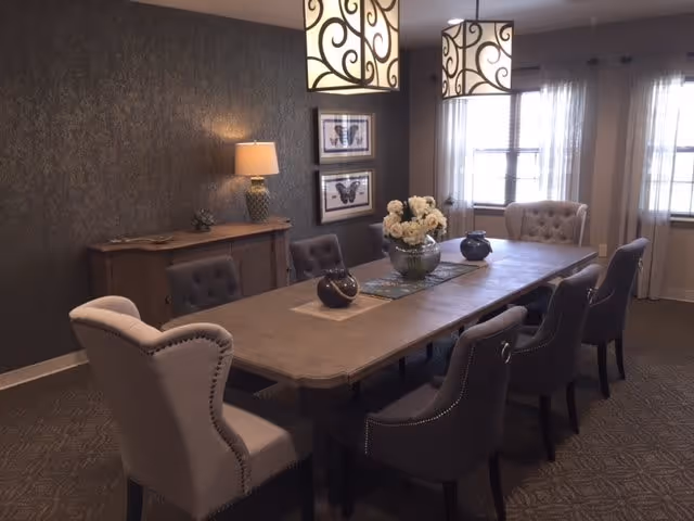 A dining room with a long rectangular wooden table surrounded by eight upholstered chairs in neutral tones. The table is decorated with a vase of white flowers and two smaller decorative items. Two pendant lights with ornate black swirl designs hang above the table. A sideboard with a lamp and decorative items is against the dark textured wall, which also features two framed butterfly artworks. Two windows with sheer curtains allow natural light into the room.
