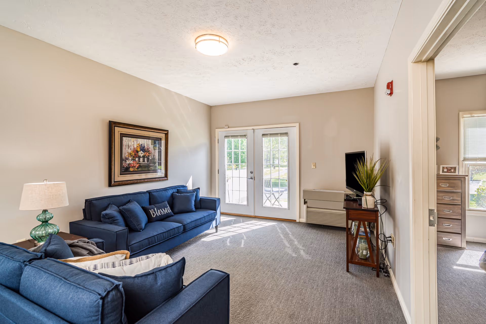 A bright living room with two blue sofas, one with a pillow that says 'Blessed'. There is a framed floral painting on the beige wall above one sofa. A glass door leads outside, letting in natural light. A small wooden table holds a TV and a plant. The room has carpeted flooring and neutral-colored walls. A doorway on the right leads to another room with a window and a chest of drawers.