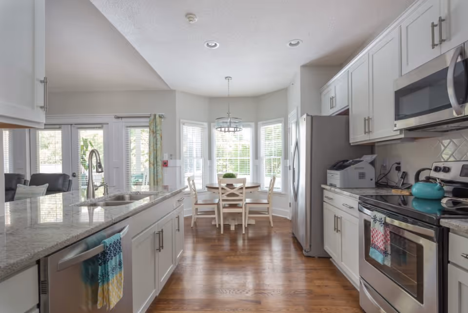 Bright kitchen with white cabinets, stainless steel appliances including a refrigerator, oven, microwave, and dishwasher. There is a kitchen island with a sink and a colorful towel hanging on the dishwasher. In the background, a round dining table with four chairs is positioned in front of large windows with white blinds, letting in natural light. Hardwood floors and a modern light fixture above the dining table complete the space.