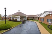 Exterior view of a single-story brick building with a covered entrance driveway, a sidewalk, and well-maintained landscaping including grass and bushes under an overcast sky.