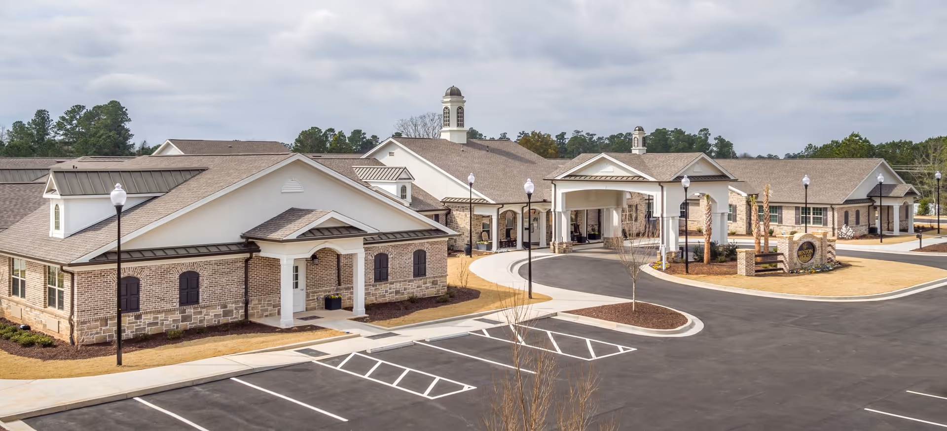 Exterior view of Benton House of Olathe, showing a large single-story building with brick and stone facade, multiple peaked roofs, a covered entrance, lamp posts, and a spacious parking lot in front under a cloudy sky.