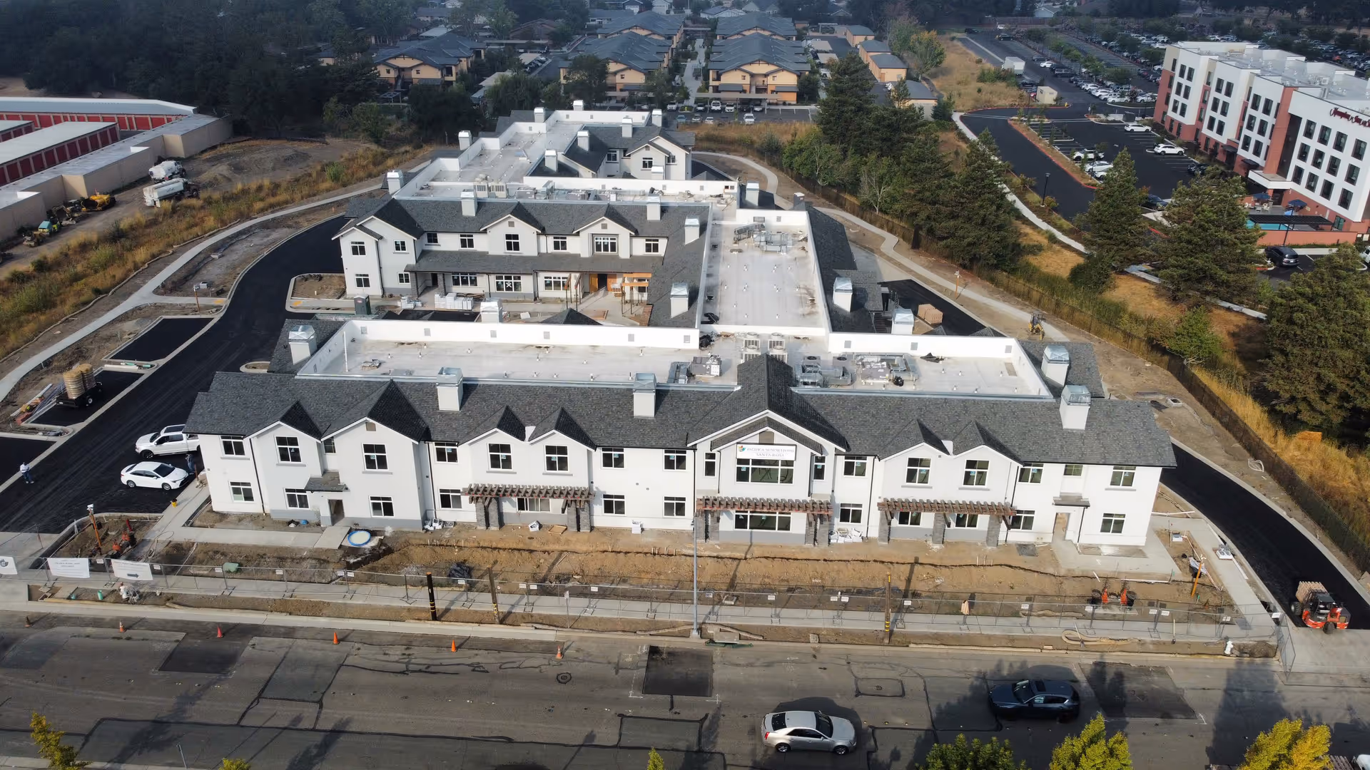 Aerial view of a large, newly constructed senior living facility building with white exterior walls and gray roofs. The building is surrounded by paved roads, parking areas, and some landscaping. There are multiple sections and entrances visible, with construction equipment and materials around the site indicating ongoing work.
