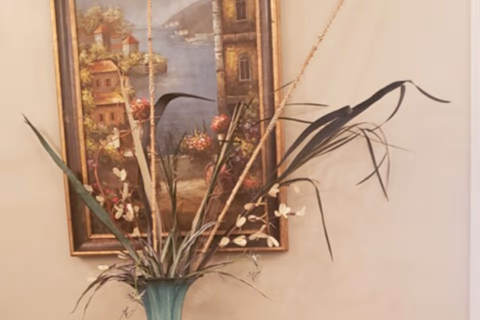 A decorative arrangement of tall dried plants and flowers in a green vase placed in front of a framed painting depicting a scenic view of buildings near water with trees and flowers.