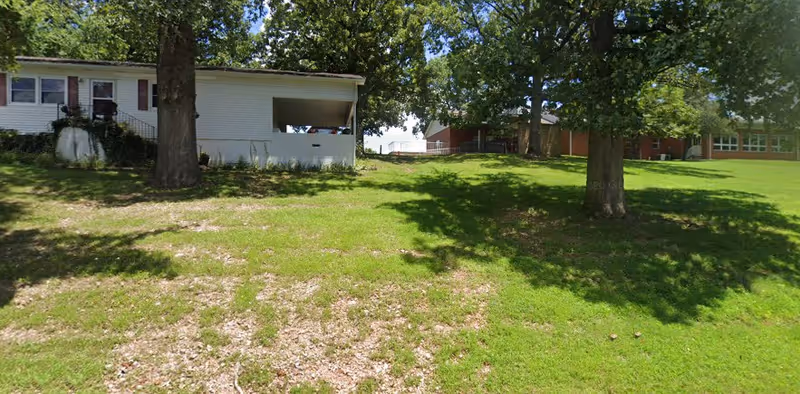 A grassy outdoor area with two large trees casting shadows. In the background, there are white and red buildings partially obscured by the trees. The sky is clear and blue.