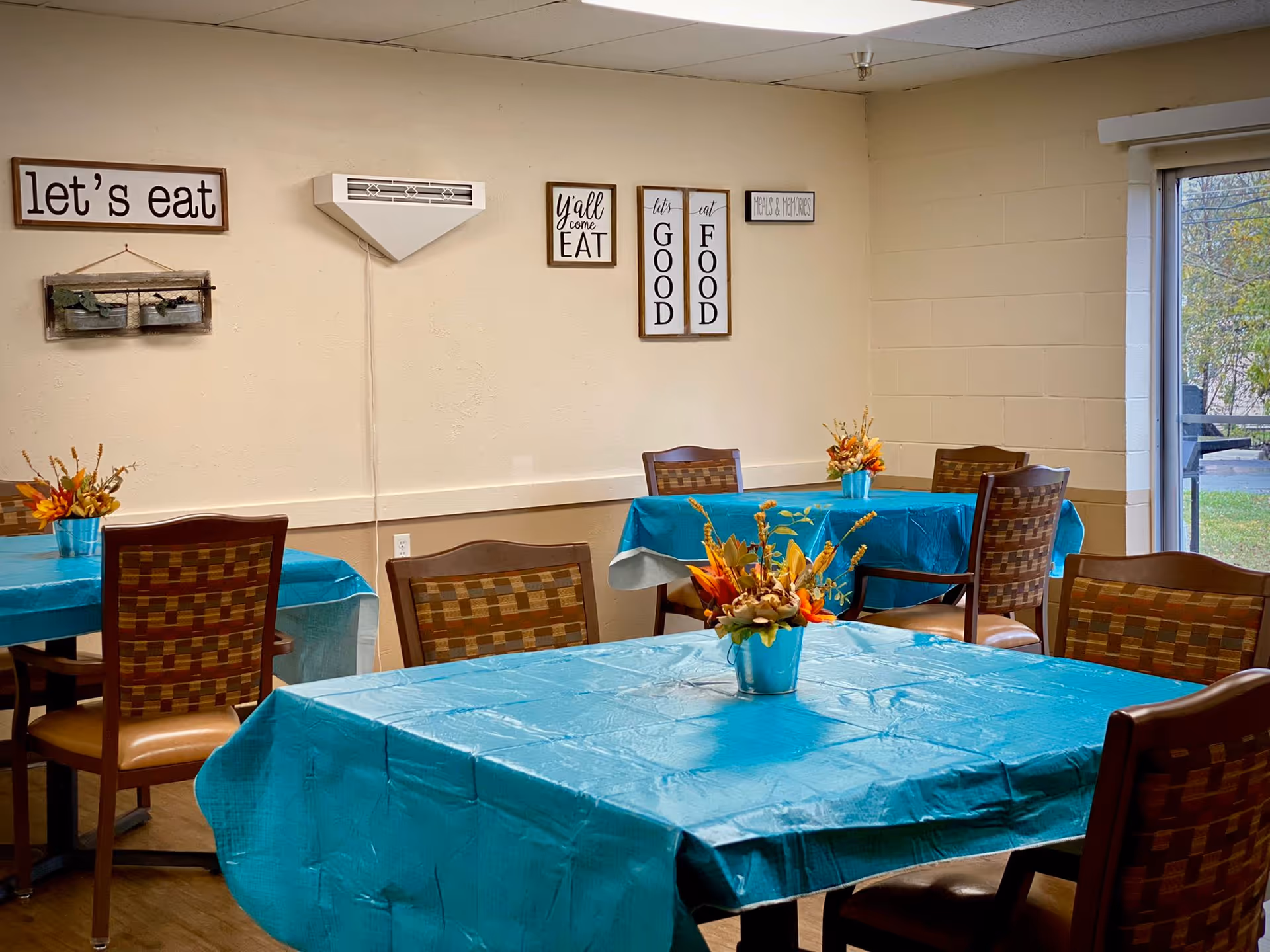 Dining area with several tables covered in blue tablecloths, each decorated with a small flower arrangement in a blue vase. The walls have multiple framed signs with phrases like 'let's eat', 'y'all come eat', 'let's eat good food', and 'peas & pumpkins'. There is a large window on the right side showing some greenery outside.