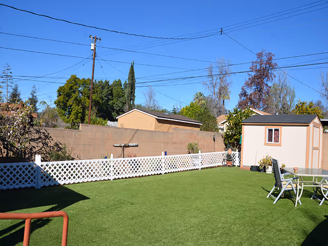 Backyard with artificial grass, a white lattice fence, a small shed, and a patio table and chairs under a clear blue sky.