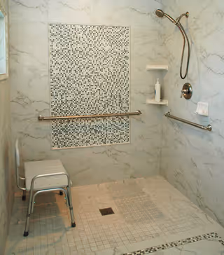 Accessible tiled walk-in shower with grab bars, a shower chair, hand-held showerhead, and a mosaic tile accent panel.