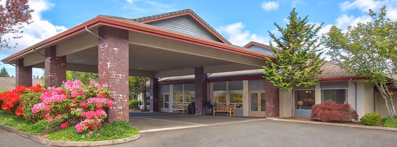 Exterior view of Alpine Way Continuing Care Community featuring a covered entrance with brick pillars, benches near the entrance, and landscaped bushes with pink and red flowers under a partly cloudy sky.