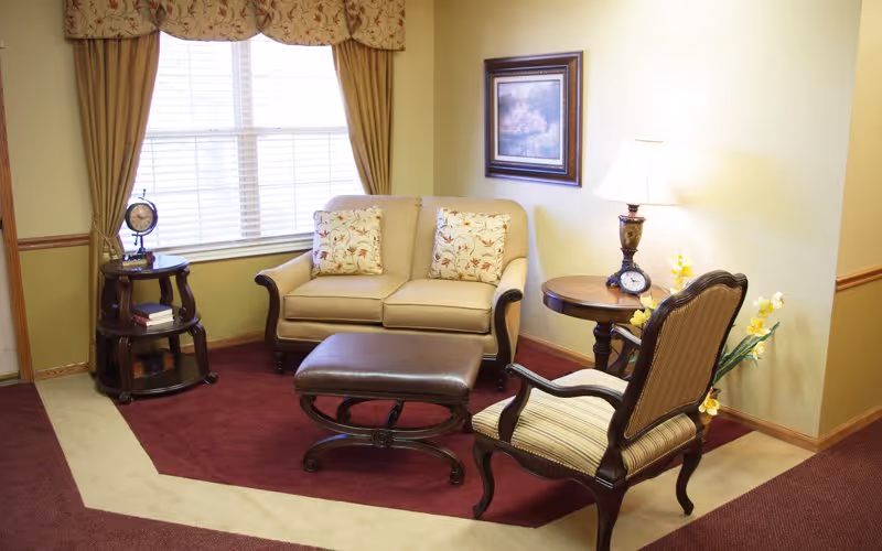 A cozy living room area with a beige loveseat adorned with two floral cushions, a striped armchair, and a brown leather ottoman on a red and beige carpet. There is a round wooden side table with a lamp and a clock, a small shelf with books and a decorative globe, a window with beige curtains, and a framed painting on the wall.