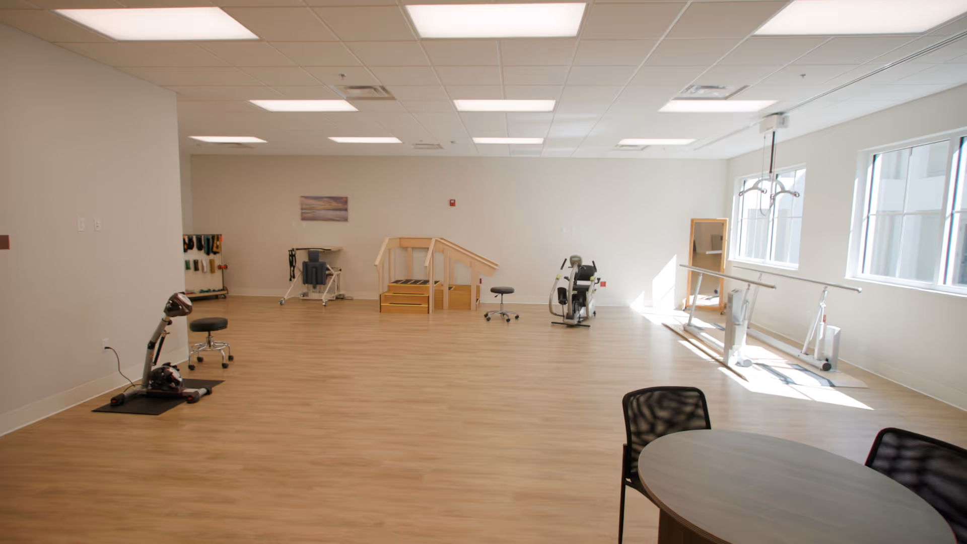 A spacious rehabilitation room with wooden flooring and white walls, equipped with exercise machines, parallel bars for walking practice, a small set of stairs with handrails, and a round table with chairs. Large windows on the right side allow natural light to fill the room.