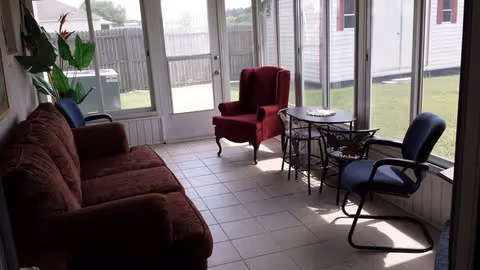 A sunlit room with large windows showing an outdoor fenced yard. Inside, there is a red upholstered armchair, a brown sofa, a blue office chair, and a small table with a decorative plate on it. There is also a plant in the corner near the sofa.