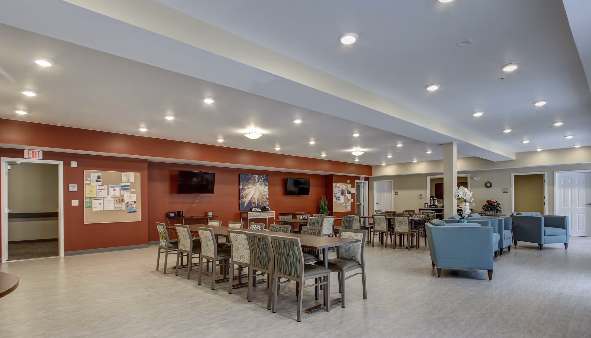 A spacious community room with multiple tables and chairs arranged for group seating. The walls are painted in warm tones with two flat-screen TVs mounted on a red accent wall. There are bulletin boards with notices and artwork on the walls. Comfortable blue armchairs are arranged in a seating area to the right. The room is well-lit with recessed ceiling lights and has a clean, modern appearance.