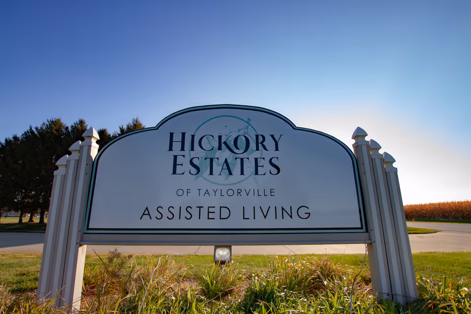 Outdoor sign for Hickory Estates of Taylorville Assisted Living, surrounded by grass and plants, with trees and a clear blue sky in the background.