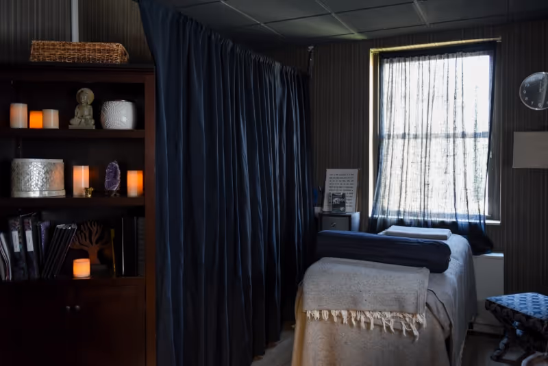 A dimly lit room with a massage table covered in beige and dark blue blankets, a window with sheer dark curtains letting in some light, a dark wooden shelf with decorative items and candles, and a dark curtain partitioning part of the room.