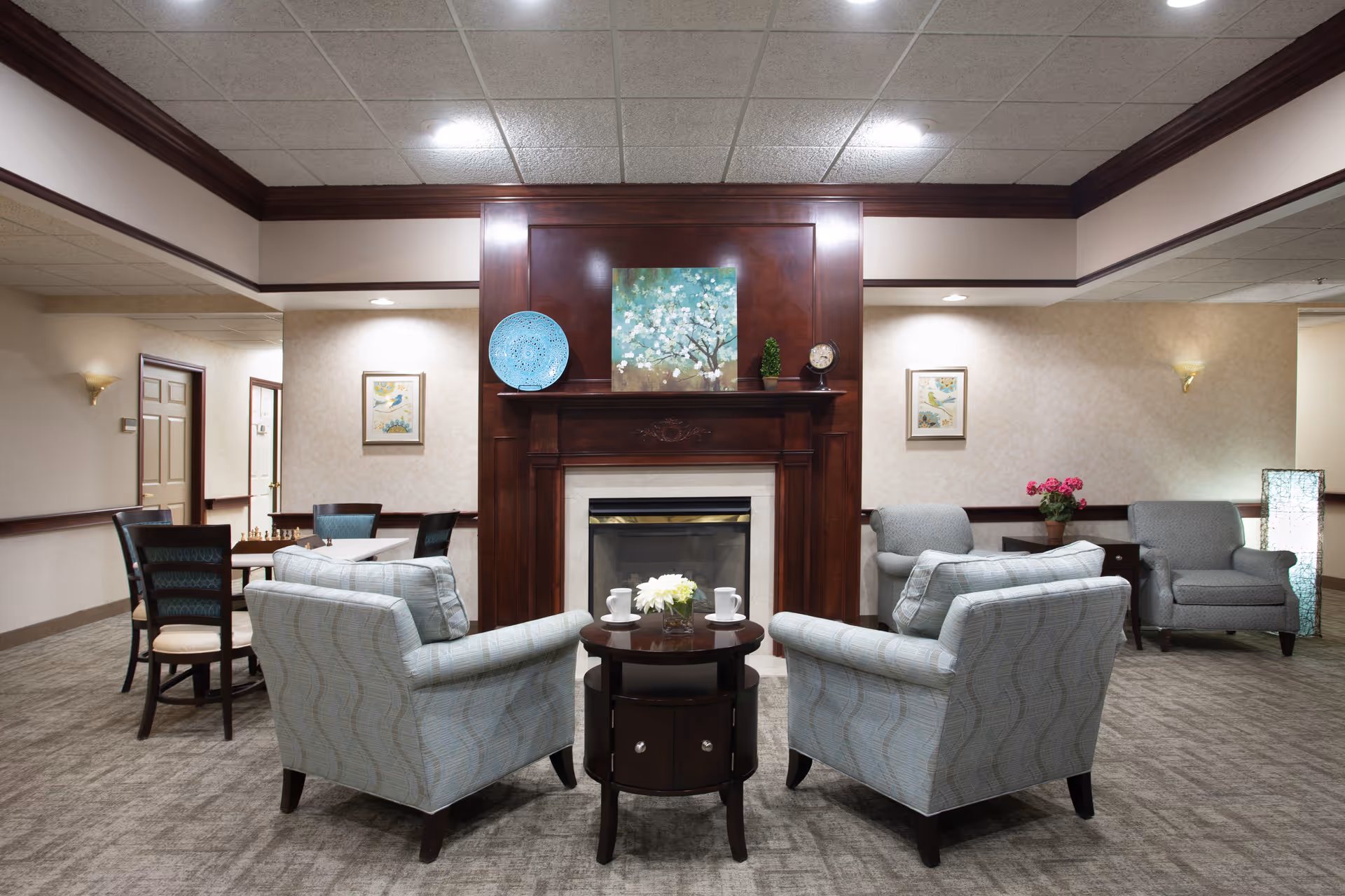 A cozy common area with a fireplace surrounded by four light blue armchairs and a small round wooden table with a flower vase and two cups. The room has beige walls with framed artwork, a carpeted floor, and additional seating including chairs and a table in the background. Decorative items such as a blue plate, a clock, and a small plant are placed on the fireplace mantel.
