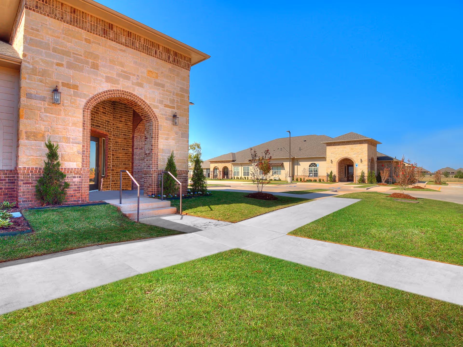 Exterior view of Iris Memory Care of Edmond showing a stone and brick building with arched entryways, manicured green lawns, small trees, and a clear blue sky.