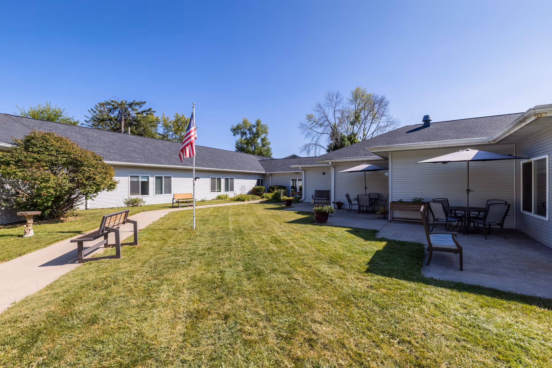 Outdoor courtyard area of Corridor Crossing Place featuring a well-maintained grassy lawn, a concrete pathway, benches, an American flag on a flagpole, patio tables with umbrellas, and a single-story building surrounding the courtyard under a clear blue sky.