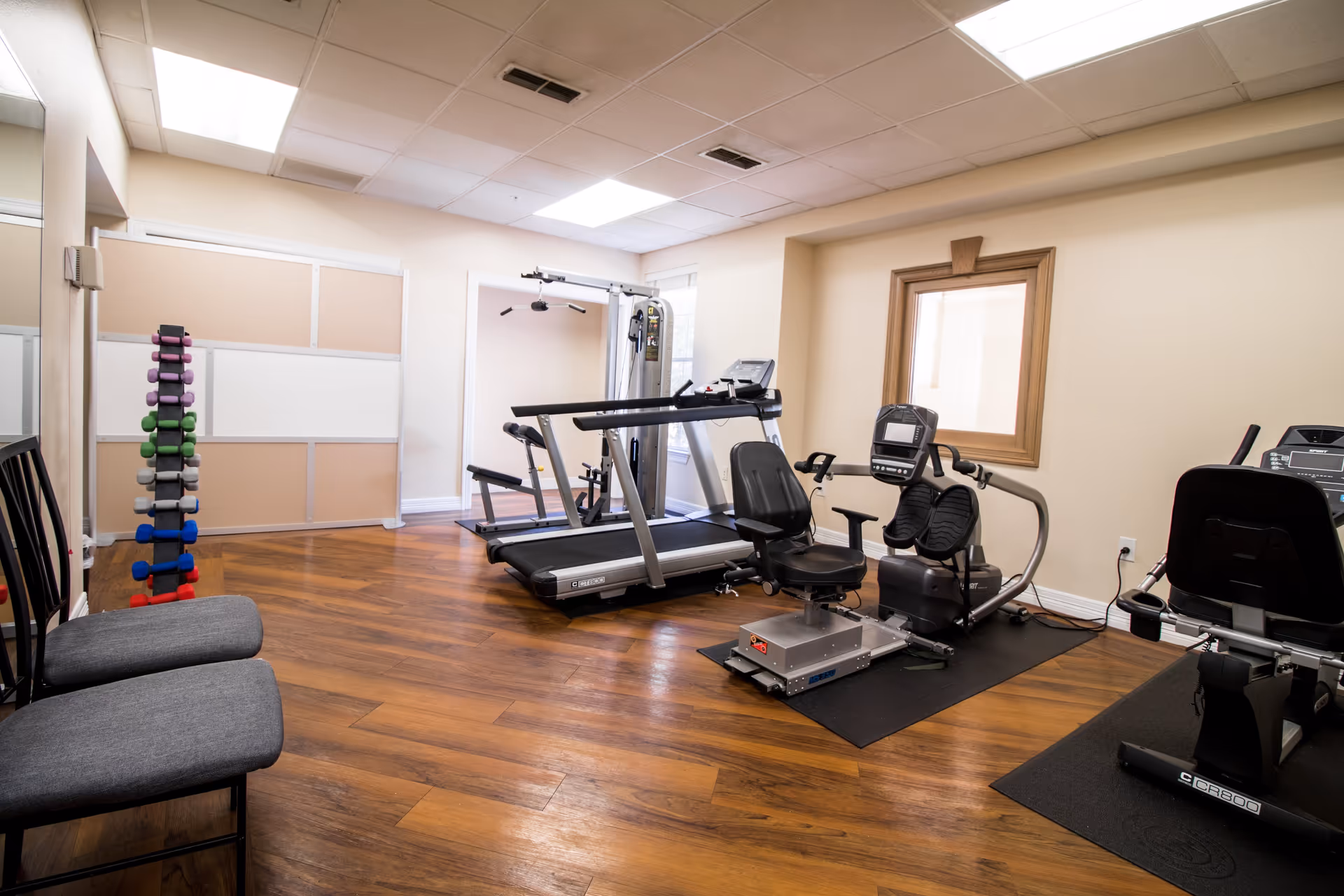 A small fitness room with wooden flooring and beige walls. The room contains exercise equipment including a treadmill, a recumbent bike, a multi-gym machine, and a rack of colorful dumbbells. There are two gray cushioned chairs on the left side and a large mirror on the left wall. A window with a wooden frame is visible on the right wall.