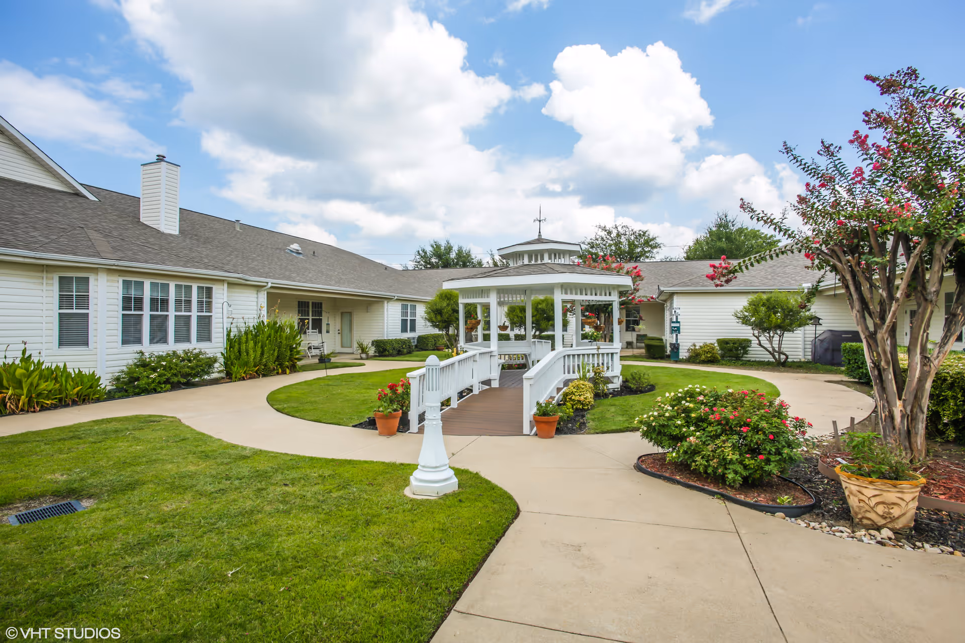 Outdoor courtyard area at Whitley Place featuring a white gazebo with a small bridge, surrounded by green grass, flowering plants, and trees. The courtyard is bordered by single-story buildings with white siding and multiple windows under a partly cloudy blue sky.