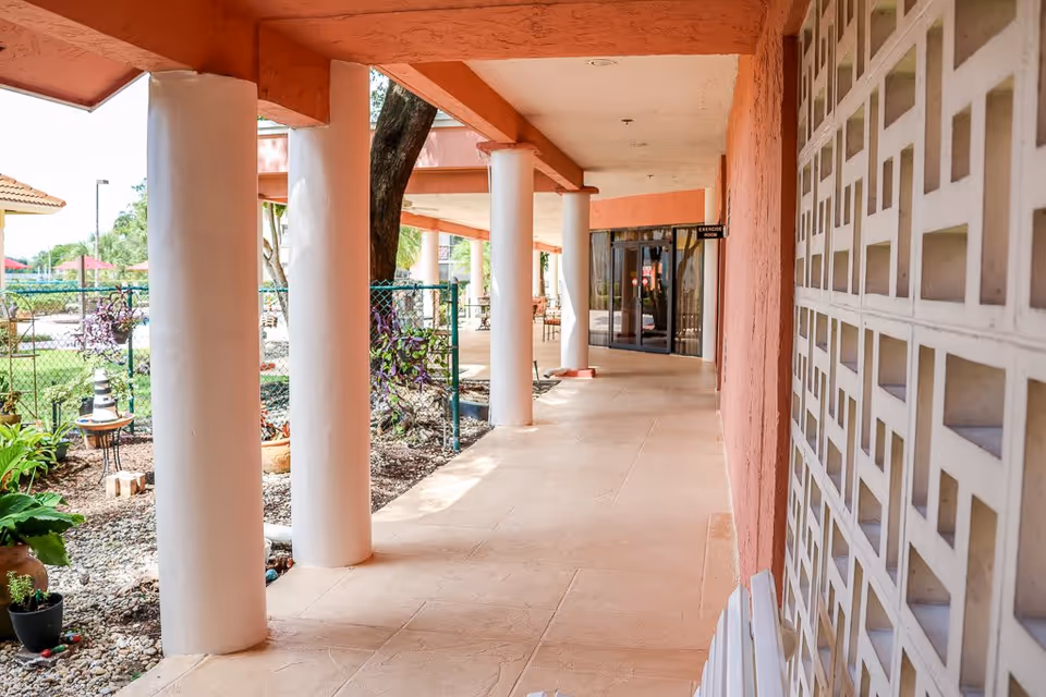 Covered outdoor walkway with white columns and a peach-colored wall on the right. There is a garden area with plants and a fence on the left side. At the end of the walkway, there is a glass door with a sign above it that reads 'Exercise Room'.