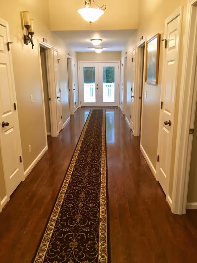 A long hallway with polished wooden floors and a dark patterned runner rug in the center. The hallway has multiple white doors on both sides, beige walls, wall-mounted candle holders, ceiling lights, and a set of double glass doors at the far end letting in natural light.