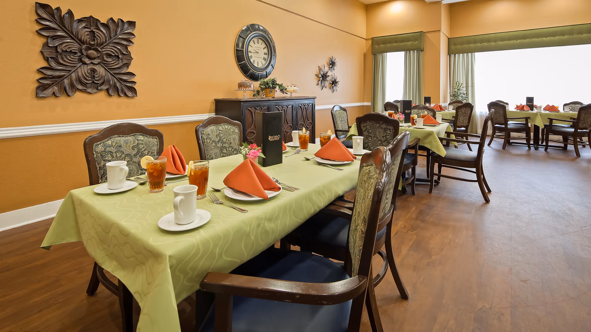 A dining room in Meadowood Assisted Living and Memory Care with tables covered in light green tablecloths, set with white plates, orange folded napkins, glasses of iced tea, and white coffee cups. The room has wooden chairs with patterned upholstery, warm yellow walls decorated with a large clock and wall art, and large windows with green curtains letting in natural light.