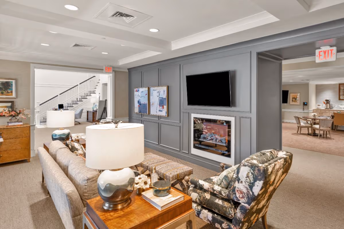 A cozy living room area in a senior living facility with a gray sofa, floral armchair, wooden side table with a lamp, and a wall-mounted TV above an electric fireplace. The room has soft lighting and neutral tones, with a view into an adjacent dining area and a staircase in the background.