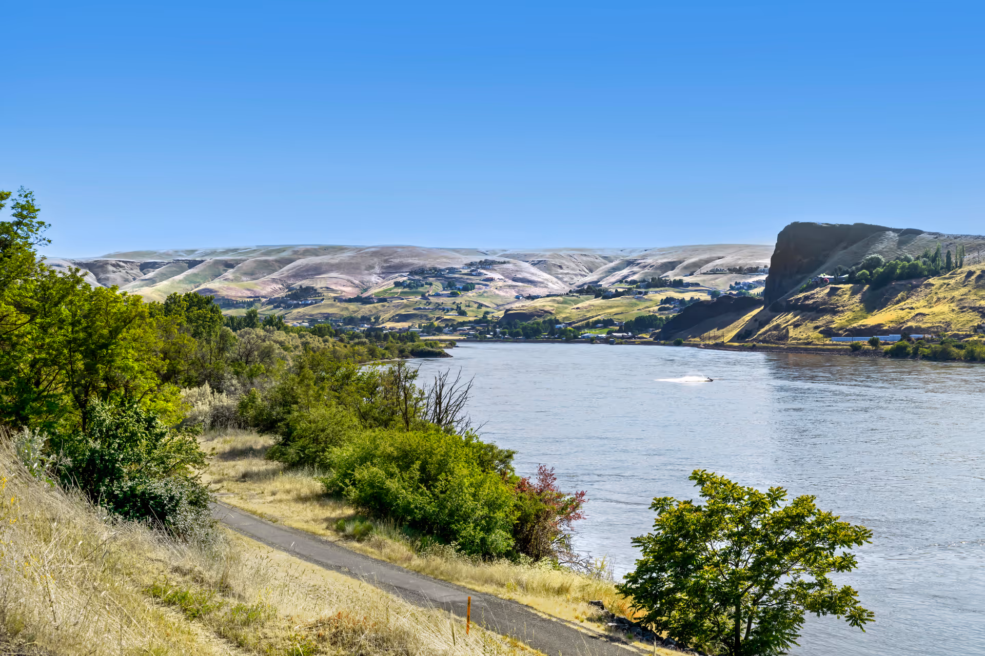 A scenic view of a river with green trees and bushes along the riverbank, hills in the background under a clear blue sky.