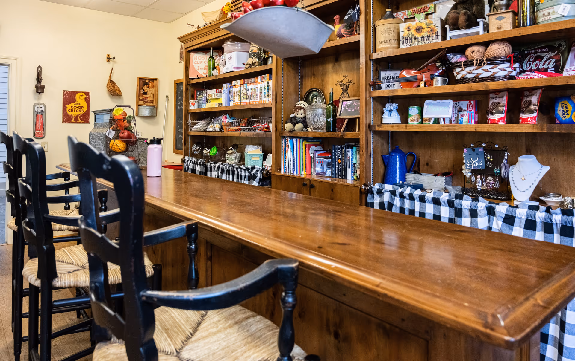 Interior view of a cozy room with a long wooden counter and four black wooden chairs with woven seats. Behind the counter is a wooden shelving unit filled with various items including books, snacks, decorative objects, and kitchenware. The room has a warm and inviting atmosphere with a mix of rustic and homey decor.