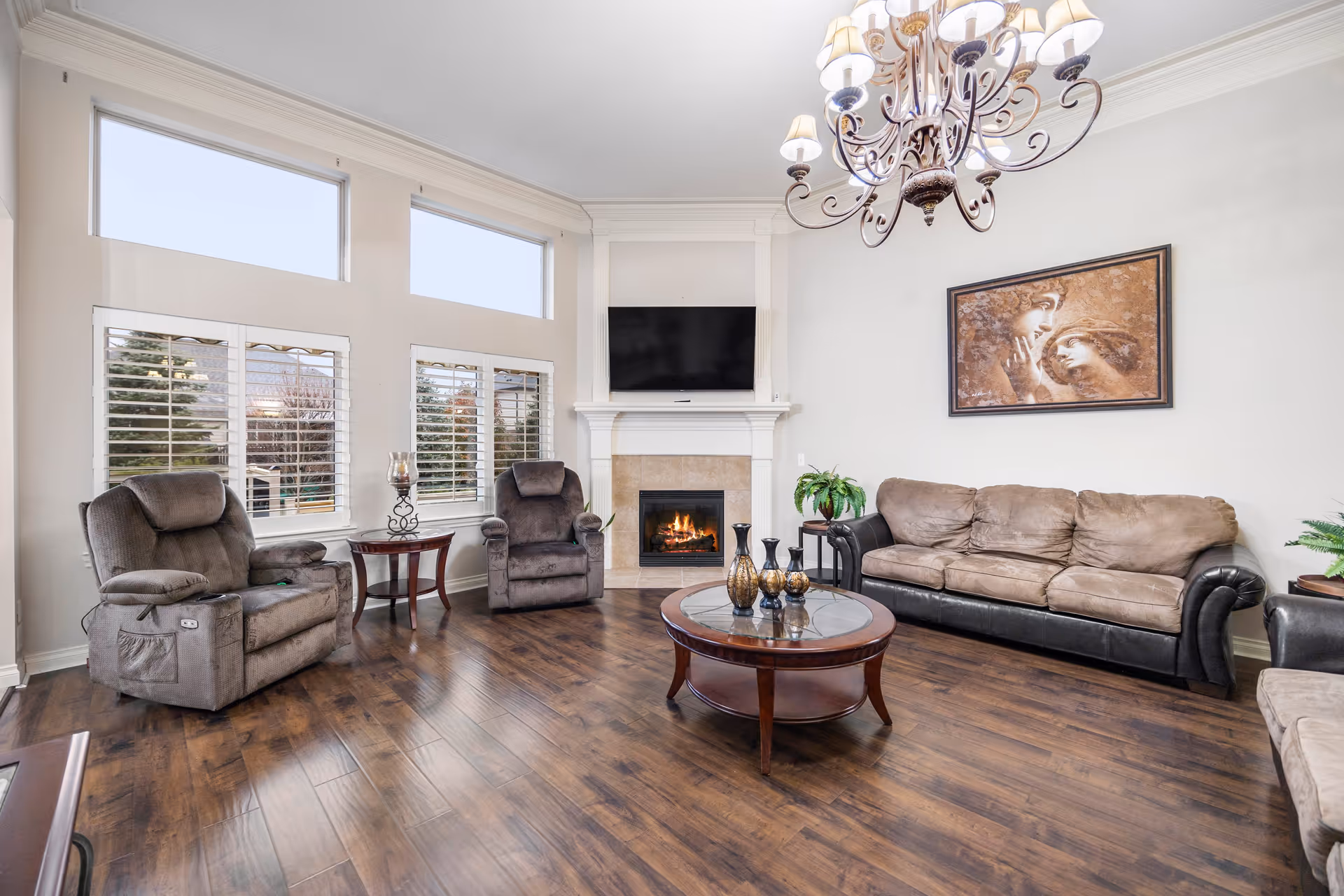 A cozy living room with dark wood flooring, two gray recliner chairs, a brown and black leather sofa, a round wooden coffee table with decorative vases, a fireplace with a mounted flat-screen TV above it, large windows with white shutters, a chandelier, and a framed artwork on the wall.