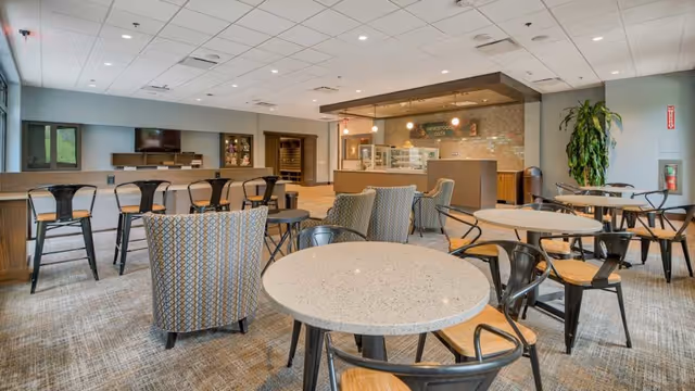 A spacious and well-lit common area with multiple round tables and chairs arranged for seating. There are cushioned armchairs and metal chairs with wooden seats. In the background, there is a counter area labeled 'Greenwood Cafe' with display cases and pendant lighting. The room has a carpeted floor, a drop ceiling with recessed lighting, and large windows letting in natural light. A tall plant and a fire extinguisher are visible near the wall.
