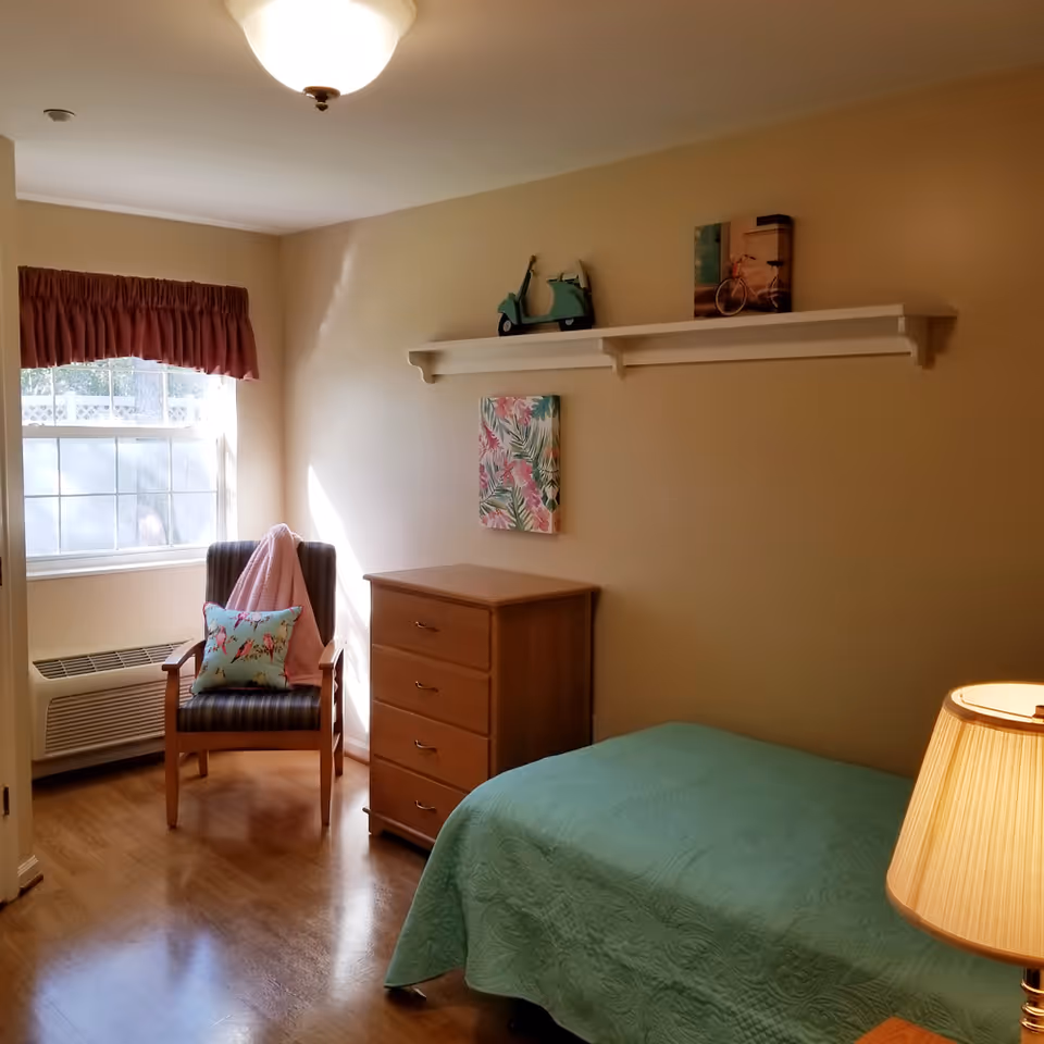 A small, cozy bedroom with a single bed covered in a green quilt. There is a wooden dresser with four drawers next to the bed. A wooden chair with a striped cushion, a pink blanket, and a decorative pillow is placed near a window with a red valance. Above the dresser, a white wall shelf holds a small green scooter model and a picture of a bicycle. A floral painting hangs on the wall beneath the shelf. A table lamp with a beige shade is partially visible on the right side.