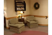 Two upholstered armchairs with a side table and lamp in a cozy sitting area with a wall shelf and wreath on the wall.