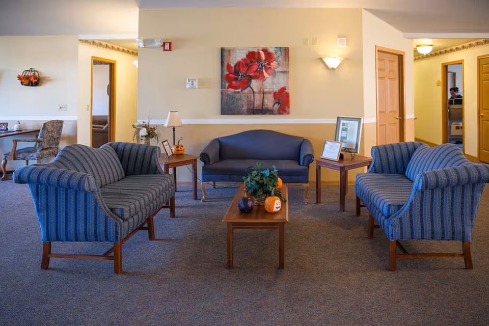 A cozy living room area in an assisted living facility with three blue upholstered sofas arranged around a wooden coffee table. The coffee table has small decorative pumpkins and a plant. There are two wooden side tables with lamps and framed pictures. A painting of red flowers hangs on the wall above the middle sofa. The room has beige walls, carpeted floor, and doorways leading to other rooms.