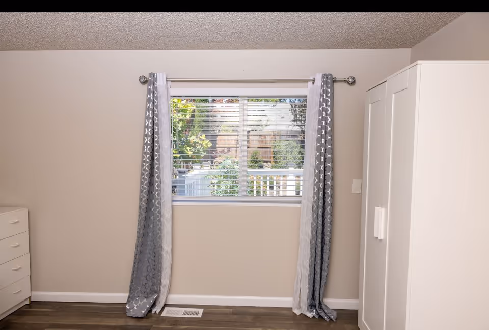 A simple bedroom interior featuring a window with gray and white patterned curtains, a white wardrobe on the right, and a white chest of drawers on the left. Outside the window, greenery and a wooden fence are visible.